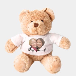May include: A light brown teddy bear wearing a white t-shirt. The t-shirt has a heart-shaped photo of two girls and the words "Best Friends". The bear has a brown nose and is made of soft material.