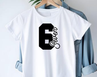 May include: White t-shirt featuring a large black number 6 and the word "Seven" in a cursive script. The garment is displayed on a wooden hanger, set against a light backdrop, with a denim jacket partially visible.