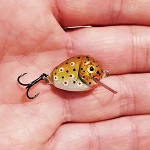 Puede incluir: Un señuelo de pesca pintado a mano con forma de pez. El señuelo tiene un cuerpo marrón con manchas negras y rojas, un anzuelo triple negro y una pequeña lengua rosa. Diseñado para atraer peces.