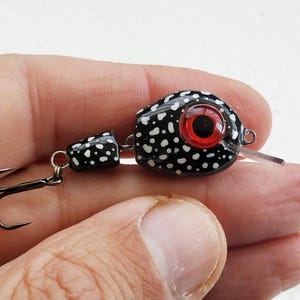 May include: A black and white fishing lure with a large red eye. The lure has a speckled pattern and a small hook attached. The lure is held in a person's hand.