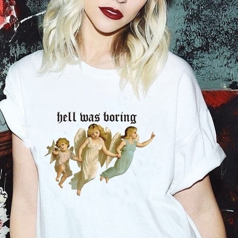 May include: White t-shirt with the text "hell was boring" printed in black. The shirt features a graphic of three angels with wings.