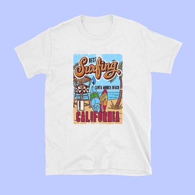 May include: White t-shirt with a vintage-style graphic design featuring the text "Best Surfing in California" with a beach scene and surfboards. Santa Monica Beach is mentioned in the design.