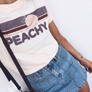 Puede incluir: Una camiseta blanca con un gr&aacute;fico de melocot&oacute;n y la palabra "Peachy" en una fuente retro. La camiseta se combina con una falda vaquera azul.