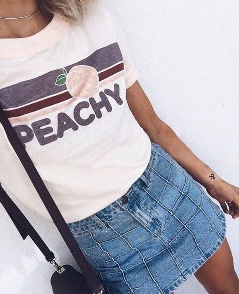 Puede incluir: Una camiseta blanca con un gr&aacute;fico de melocot&oacute;n y la palabra "Peachy" en una fuente retro. La camiseta se combina con una falda vaquera azul.