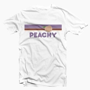 Puede incluir: Camiseta blanca con un gr&aacute;fico de melocot&oacute;n y la palabra "PEACHY" en texto morado.