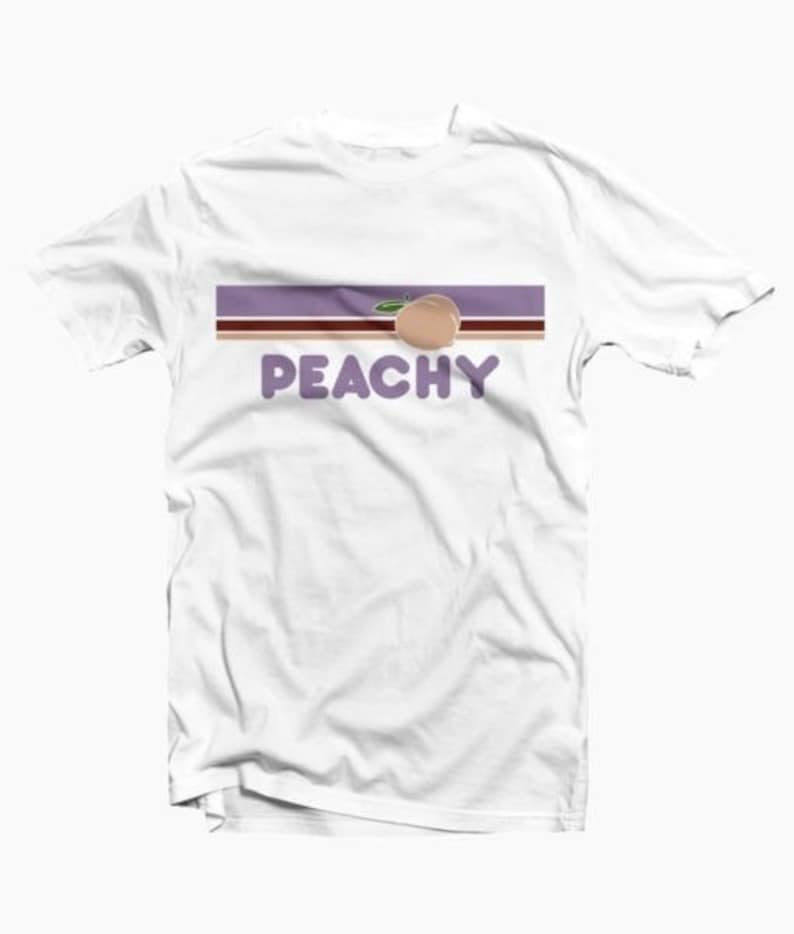 Puede incluir: Camiseta blanca con un gr&aacute;fico de melocot&oacute;n y la palabra "PEACHY" en texto morado.