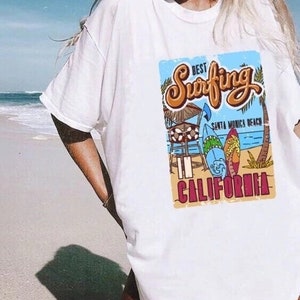 May include: White t-shirt with a graphic print of a beach scene with surfboards and the text "Best Surfing in California" with the words "Santa Monica Beach" underneath.
