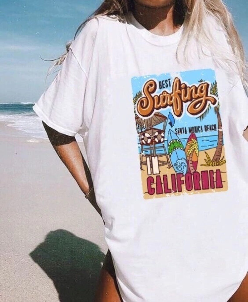 May include: White t-shirt with a graphic print of a beach scene with surfboards and the text "Best Surfing in California" with the words "Santa Monica Beach" underneath.
