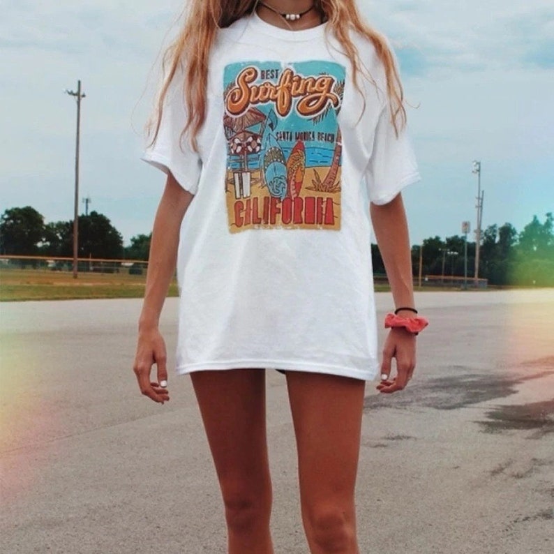 May include: White t-shirt with a graphic print of a beach scene and the text "Best Surfing Santa Monica Beach California".