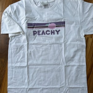 Puede incluir: Camiseta blanca con un dise&ntilde;o de rayas moradas y negras y la palabra "PEACHY" en letras moradas. Un melocot&oacute;n rosa con un tallo verde est&aacute; por encima de la palabra.