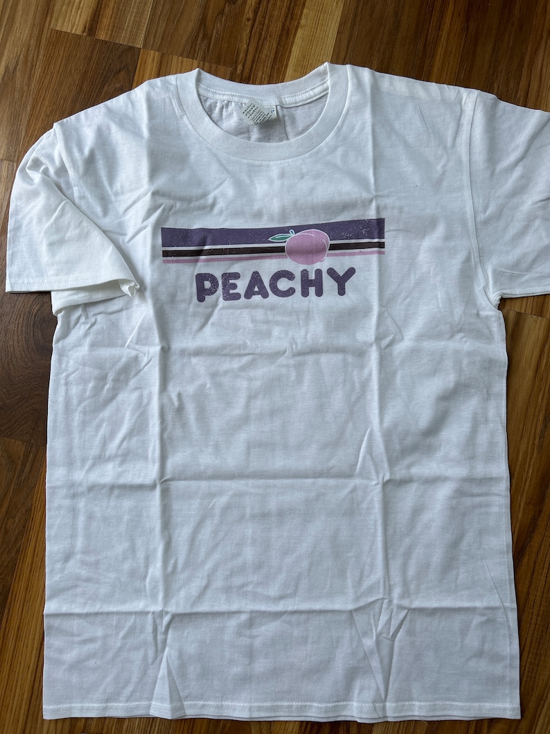 Puede incluir: Camiseta blanca con un dise&ntilde;o de rayas moradas y negras y la palabra "PEACHY" en letras moradas. Un melocot&oacute;n rosa con un tallo verde est&aacute; por encima de la palabra.