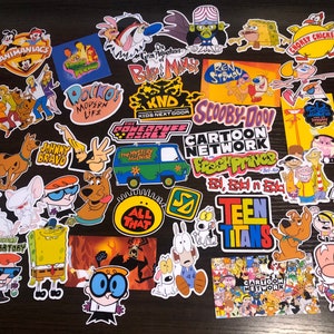 Puede incluir: Una colección de pegatinas coloridas de personajes de dibujos animados de series populares. Las pegatinas incluyen personajes de Animaniacs, Scooby-Doo, el Laboratorio de Dexter, y más. Las pegatinas están dispuestas sobre una superficie oscura.