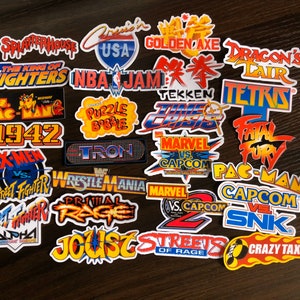 May include: A selection of vibrant stickers showcasing logos from retro arcade games. The stickers include titles such as "Pac-Man," "Street Fighter," "Marvel vs. Capcom," and "NBA Jam." The stickers are displayed on a dark wooden surface.