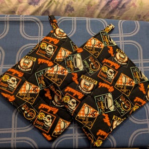 May include: Two black quilted pot holders with a coffee-themed pattern. The design includes coffee cups, stamps, and text like "Coffee Shop" and "Robust Coffee." Each pot holder has a small loop for hanging.
