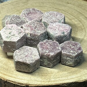 May include: A group of nine pink ruby crystals, each with a hexagonal shape, are displayed on a wooden surface. The crystals are rough and have a natural, unpolished appearance.