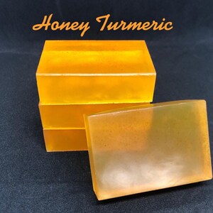 May include: Four rectangular bars of soap are stacked and arranged on a dark surface. The soap is a translucent, golden yellow color, with visible flecks throughout. The text "Honey Turmeric" is written in orange script at the top.