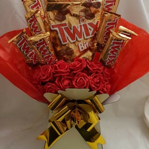 Puede incluir: Un ramo de rosas rojas con un lazo dorado y una variedad de barras de chocolate Twix en envoltorios dorados.
