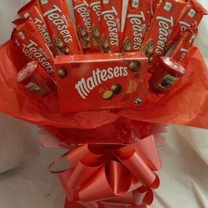 Puede incluir: Una cesta de regalo roja llena de barras de chocolate y velas. Las barras de chocolate están envueltas en papel de aluminio rojo y tienen impresas las palabras "Maltesers Teasers". La cesta está atada con una cinta roja y tiene un lazo rojo en la parte superior. La cesta también incluye una caja de chocolates Maltesers.