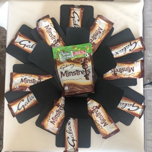 May include: A black and white explosion box filled with chocolate bars. The box is decorated with black cardstock and features a variety of Galaxy and Minstrels chocolate bars. The box also includes a small green and white bag with the text "WIN £20,000" and "Galaxy Minstrels".