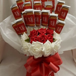 May include: A bouquet of red and white roses with a red bow, arranged in a red box. The bouquet is surrounded by white tissue paper and decorated with individually wrapped Lotus Biscoff biscuits.