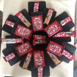 Może przedstawiać: Czarne i czerwone pudełko eksplodujące wypełnione czekoladowymi batonikami Kit Kat. Pudełko jest ozdobione czarnym papierem, a batoniki są ułożone w okrągły wzór.