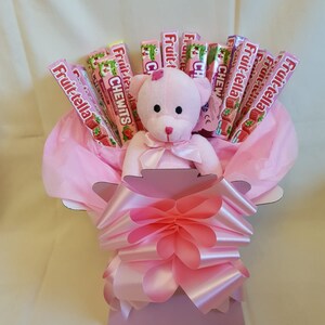 May include: Pink teddy bear sitting in a pink gift basket with pink tissue paper and a pink bow. The basket is filled with strawberry flavored Fruit-tella candy.
