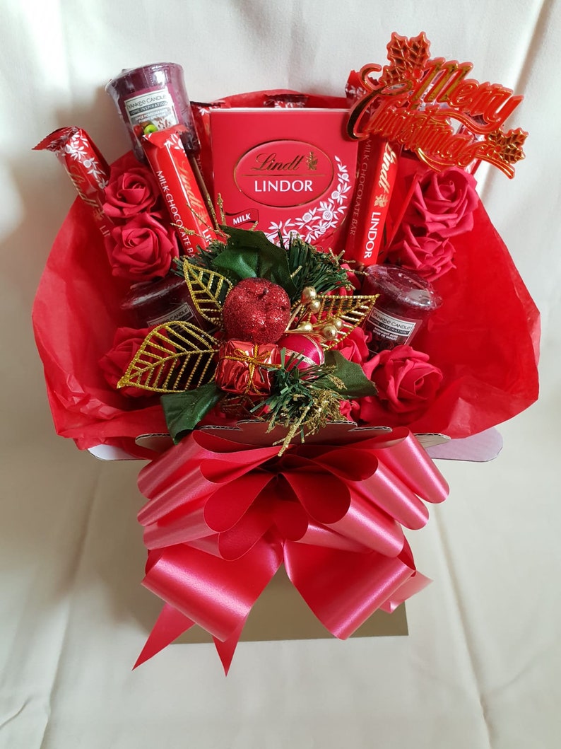 Puede incluir: Una cesta de regalo roja llena de chocolate Lindor, rosas rojas, velas y un letrero que dice "Feliz Navidad". La cesta est&aacute; atada con un lazo rojo.