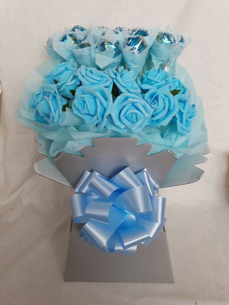 Op de afbeelding: Een boeket van lichtblauwe kunstrozen met een blauw lint en een middenstuk gevuld met verpakte chocoladebonbons.