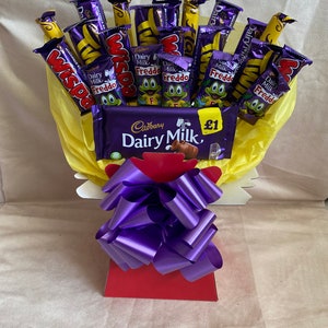May include: A yellow and white gift basket filled with chocolate bars. The basket includes Cadbury Dairy Milk, Wispa, and Freddo chocolate bars. The basket is decorated with a purple bow and a £1 price tag.