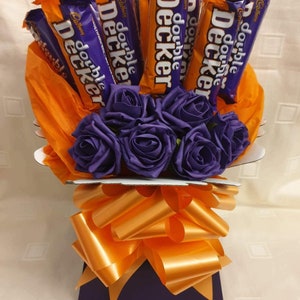 May include: A bouquet of purple silk roses arranged in a purple and orange gift box with a large orange bow. The bouquet is surrounded by orange tissue paper and topped with a dozen Double Decker chocolate bars.