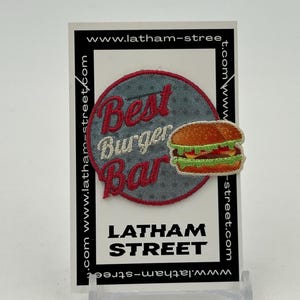 May include: An embroidered patch featuring the text "Best Burger Bar" in red, white, and blue, with a cartoon burger. The patch is displayed on a white card with the website address "www.latham-street.com" printed on it.