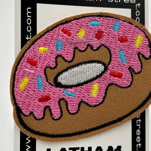 May include: An embroidered donut patch in pink and brown with colourful sprinkles. The donut has a white centre and a brown outline.