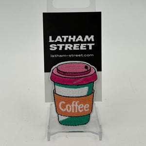 May include: An embroidered patch featuring a coffee cup in pink, green and orange colours. The word "Coffee" is embroidered on the cup. The patch is displayed on a black card with the text "Latham Street" and "latham-street.com" printed on it.