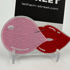 May include: A pink and red embroidered patch of a bubblegum popping with a red lip. The patch is on a white stand. The text "latham-street.com" is visible in the background.