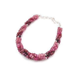 May include: A pink and burgundy beaded bracelet with a silver clasp. The bracelet is made of small, faceted beads in varying shades of pink and burgundy. The beads are strung together in a twisted design.