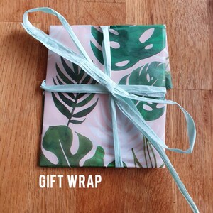 May include: A gift wrapped in pink and green tropical leaf patterned paper with a light blue ribbon tied in a bow. The text "GIFT WRAP" is printed on the bottom of the image.