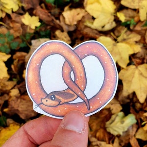 May include: A brown and white pretzel shaped sticker with a snake wrapped around it. The snake has a brown body with white spots and black eyes.