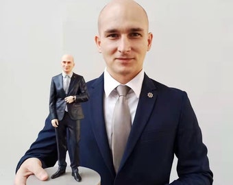 May include: A man in a dark blue suit holds a small 3D printed figurine of himself, also wearing a dark blue suit. The figurine is standing on a round grey base.