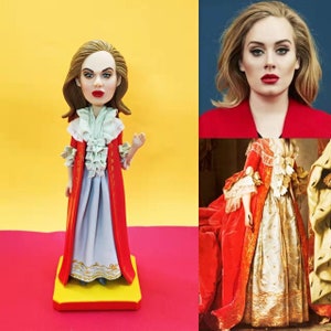 Könnte beinhalten: Eine Bobblehead-Figur einer Frau mit braunen Haaren, die ein rot-weißes Kleid trägt. Die Figur steht auf einem gelben und roten Sockel. Die Frau trägt ein rot-weißes Kleid mit einem weißen Spitzenkragen. Die Figur ist eine Nachbildung einer berühmten Sängerin.