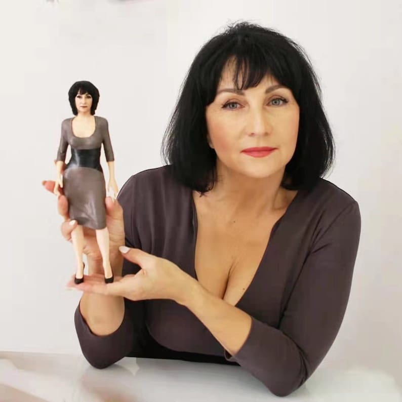 May include: A woman holds a 3D printed figurine of a woman wearing a brown dress and black heels. The figurine is about 15 cm tall. The woman holding the figurine is wearing a brown top with a deep V-neck.