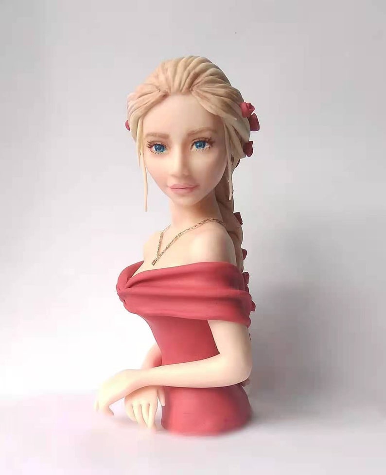 K&ouml;nnte beinhalten: Eine Skulptur eines Frauenbuste mit langen blonden Haaren, die ein rotes, schulterfreies Kleid und eine goldene Halskette tr&auml;gt. Sie hat blaue Augen und einen zarten Gesichtsausdruck.