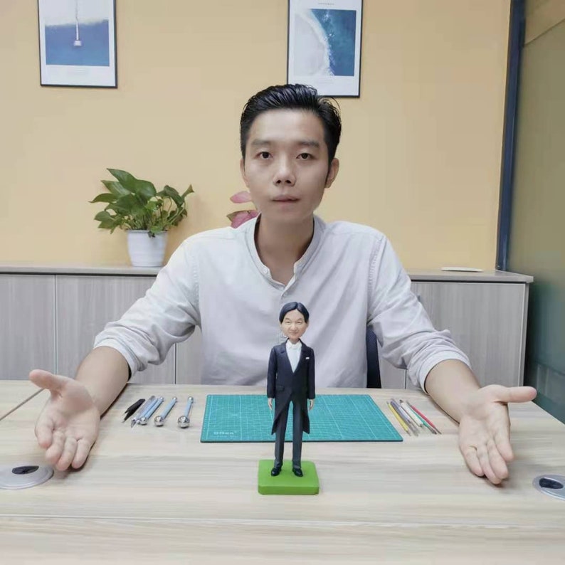 May include: A person is holding out their hands on either side of a small figurine of a person wearing a black suit and a white shirt. The figurine is standing on a green base. The person is sitting at a table with a green cutting mat and tools.