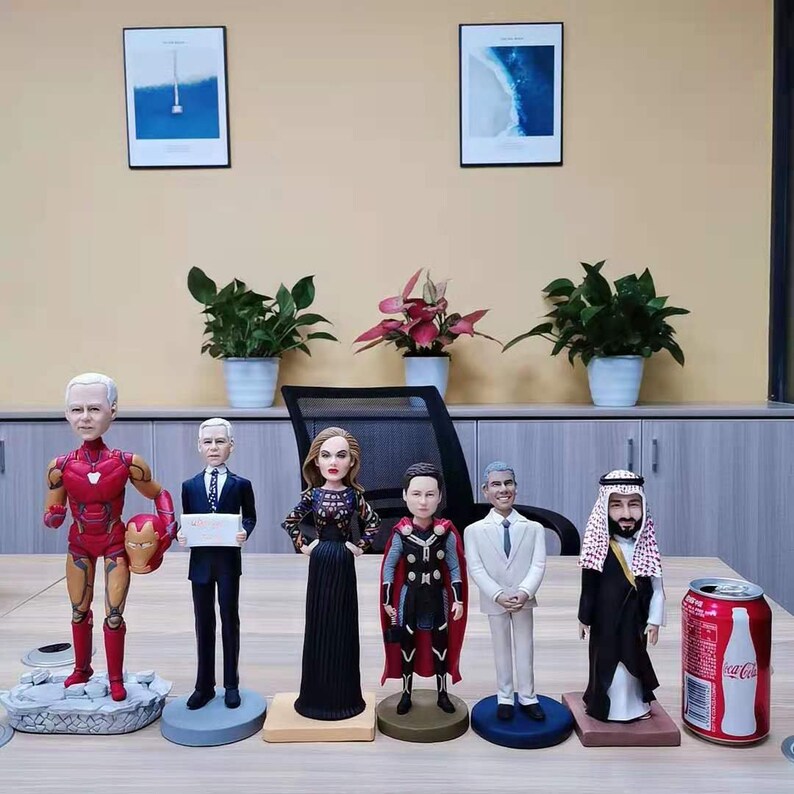 May include: A group of seven custom-made bobbleheads, each with a unique design and personality. The bobbleheads are arranged on a table in front of a wall with two framed pictures. The bobbleheads include a person dressed as Iron Man, a person in a suit, a woman in a black dress, a person dressed as Thor, a person in a white suit, a person in a white robe with a red and white headdress, and a can of Coca-Cola.
