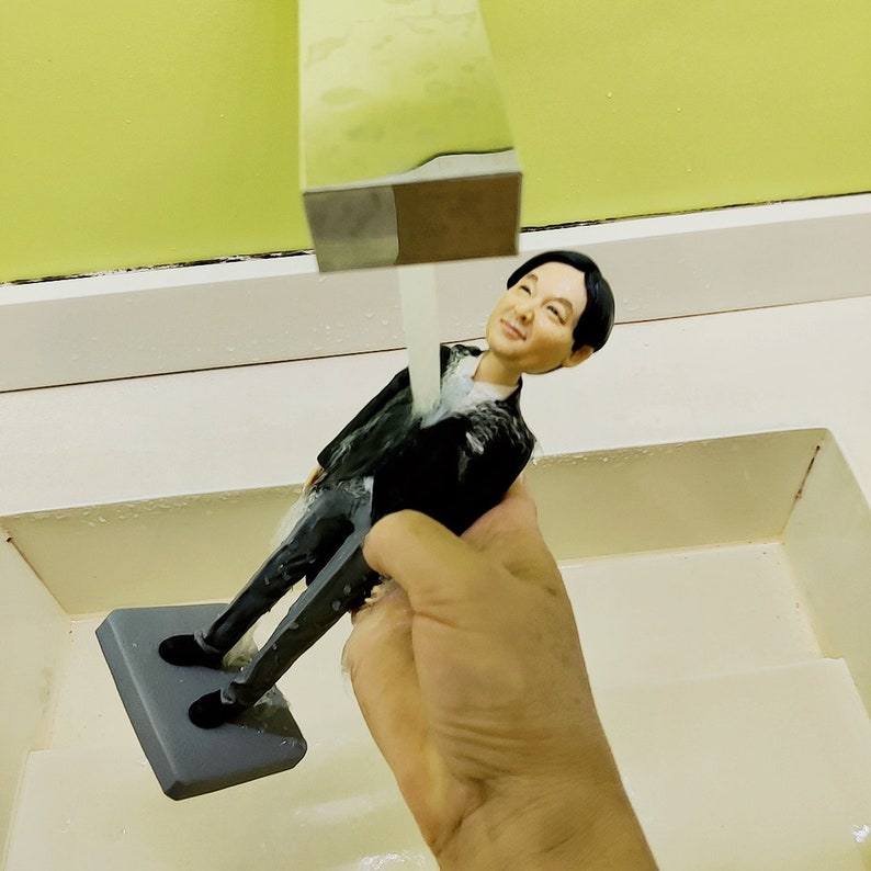 May include: A 3D printed figurine of a person wearing a black suit and white shirt is being rinsed under a faucet in a sink. The figurine is standing on a black base.