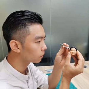 May include: A person is working on a small, detailed clay sculpture of a man's head. The sculpture is realistic and has a brown complexion. The person is using a small tool to shape the clay.