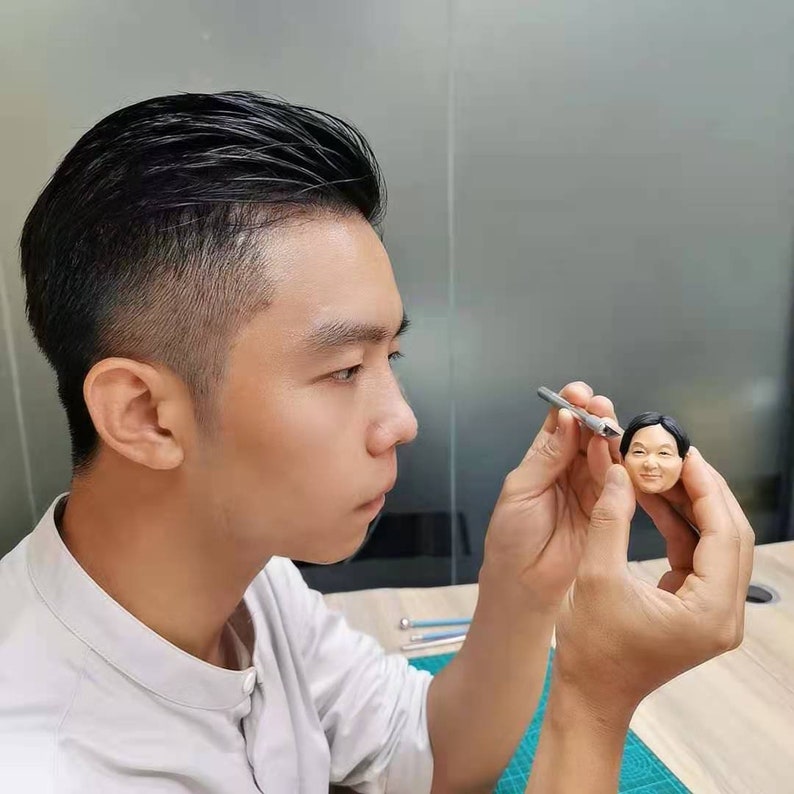 May include: A person is working on a small, detailed clay sculpture of a man's head. The sculpture is realistic and has a brown complexion. The person is using a small tool to shape the clay.