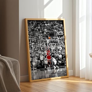 May include: A framed vertical wall art print featuring a black and white photograph of a basketball game in progress. A player in a red jersey is jumping to shoot an orange basketball toward a hoop, surrounded by a large crowd of spectators.
