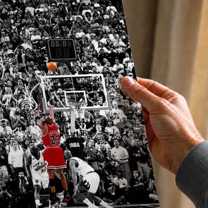 May include: A black and white photograph print featuring a basketball player in a red jersey with the number 23 jumping to shoot a basketball toward a hoop. The background shows a crowded arena with spectators and a digital scoreboard.