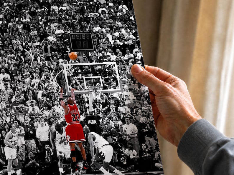 May include: A black and white photograph print featuring a basketball player in a red jersey with the number 23 jumping to shoot a basketball toward a hoop. The background shows a crowded arena with spectators and a digital scoreboard.