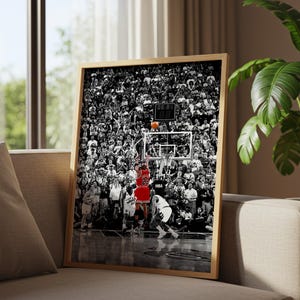 May include: A framed vertical wall art print featuring a black and white photograph of a crowded basketball arena. A single player in a red jersey is jumping to shoot an orange basketball toward the hoop, standing out against the monochrome crowd.